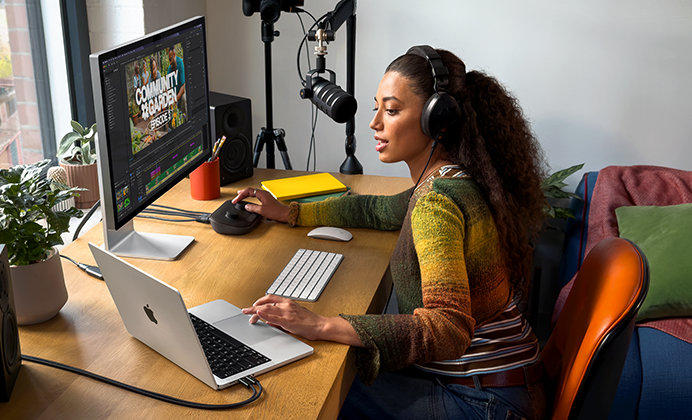 Žena sedí doma u pracovního stolu a používá MacBook Pro připojený k externímu monitoru, má nasazená sluchátka a mluví do mikrofonu