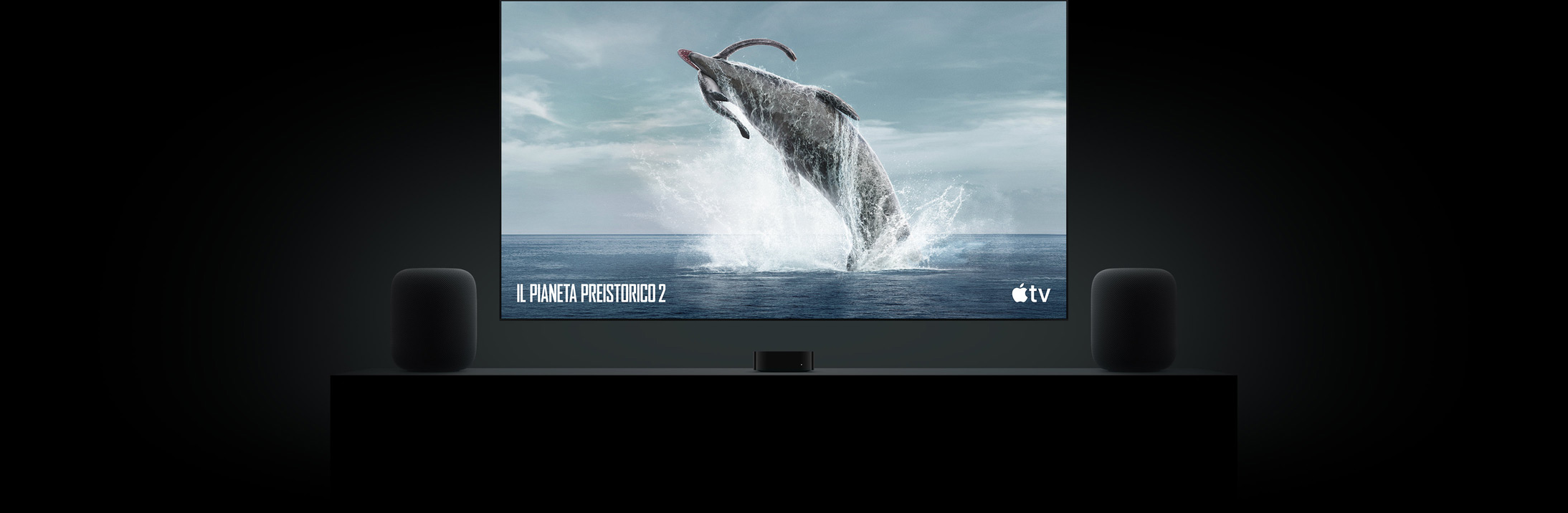 Un grande televisore a schermo piatto che mostra una vivida immagine di un dinosauro, tratta dalla docuserie Il Pianeta preistorico. Il televisore è appeso sopra una Apple TV 4K e ai lati ci sono due altoparlanti HomePod appoggiati su un mobile da soggiorno.