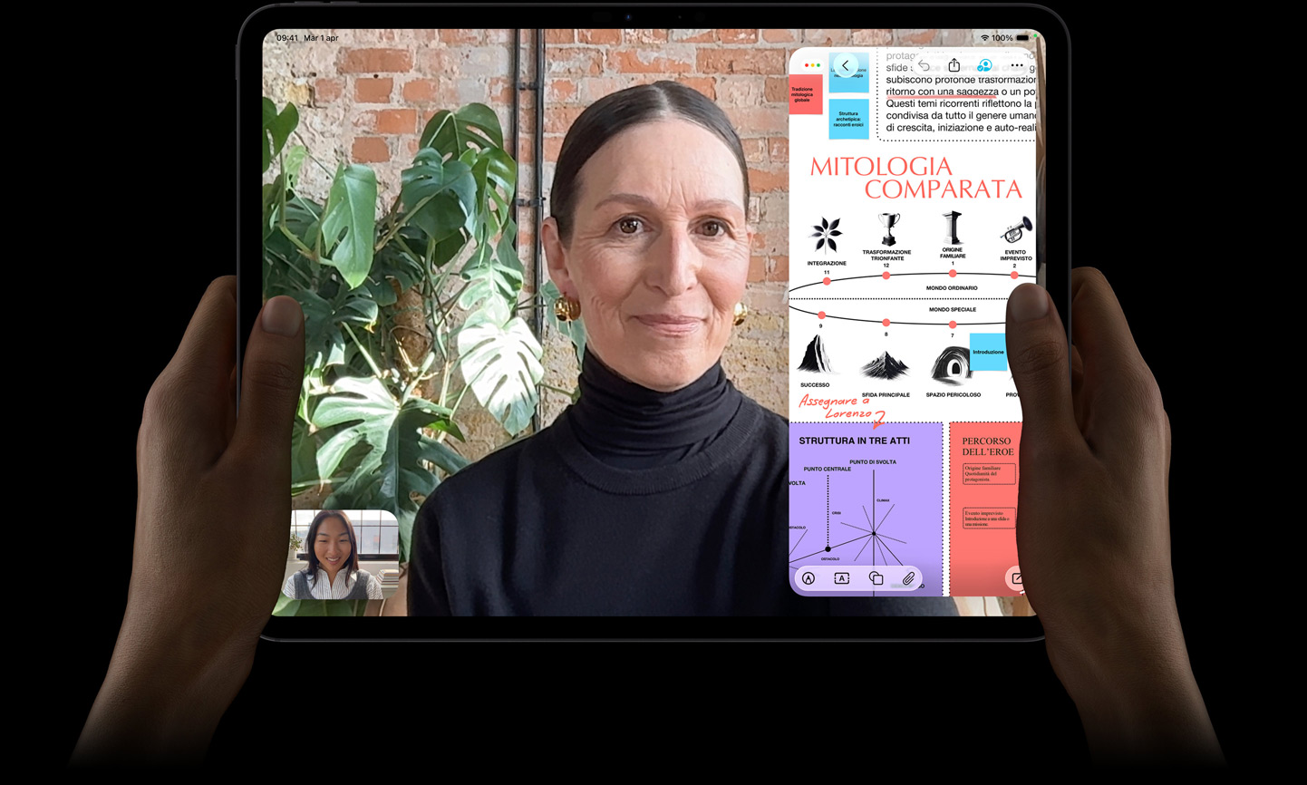 Un iPad Pro nero siderale, mostrato di fronte, tenuto fra due mani in orizzontale, angoli arrotondati, cornice del display nera, display che mostra una conversazione FaceTime con traduzione automatica in tempo reale, e un’app didattica con una presentazione sulla mitologia comparata con testo, diagrammi e note scritte a mano