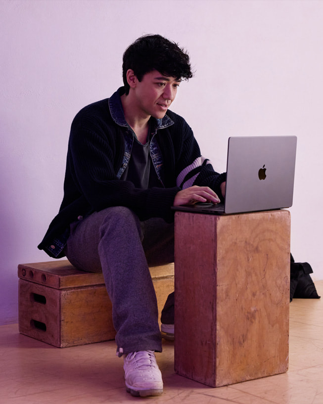 Kyle Hanagami sits on a crate in a studio setting, typing on MacBook Pro that’s placed on another crate.