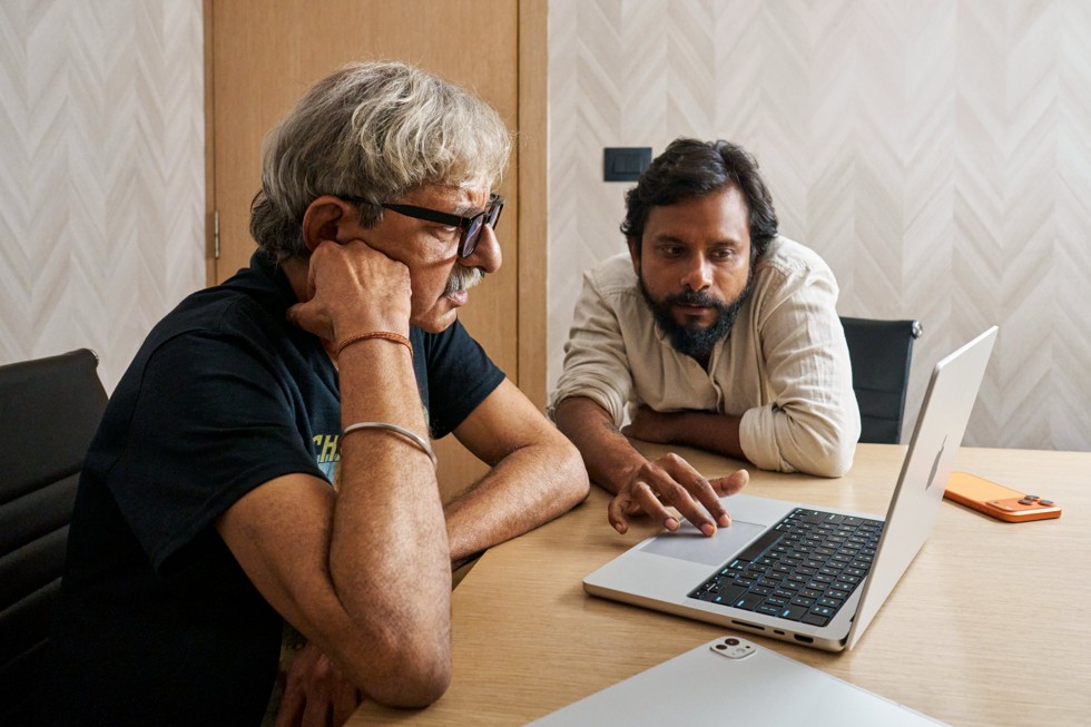 Ritesh Sharma werkt samen met een van de leden van de crew aan een bureau met daarop een MacBook Pro en een iPhone 17 Pro Max.