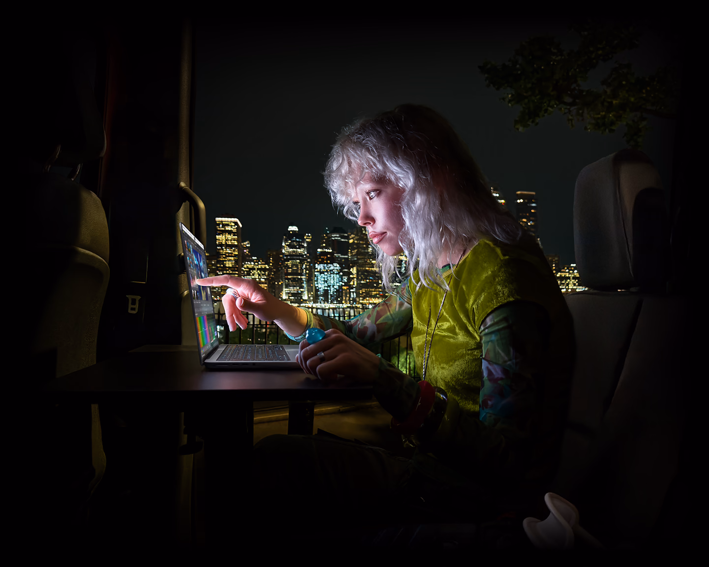 A person sitting in the back seat of a van, the open passenger door reveals a city skyline at night, MacBook Pro with PROLED XDR Display on a worktable, the person is illuminated by the screen while using the Touch feature to edit a video in DaVinci Resolve Studio