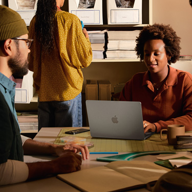 Des collègues en train de collaborer autour d’un bureau où sont étalés différents outils de création graphique. L’une des personnes utilise un MacBook.