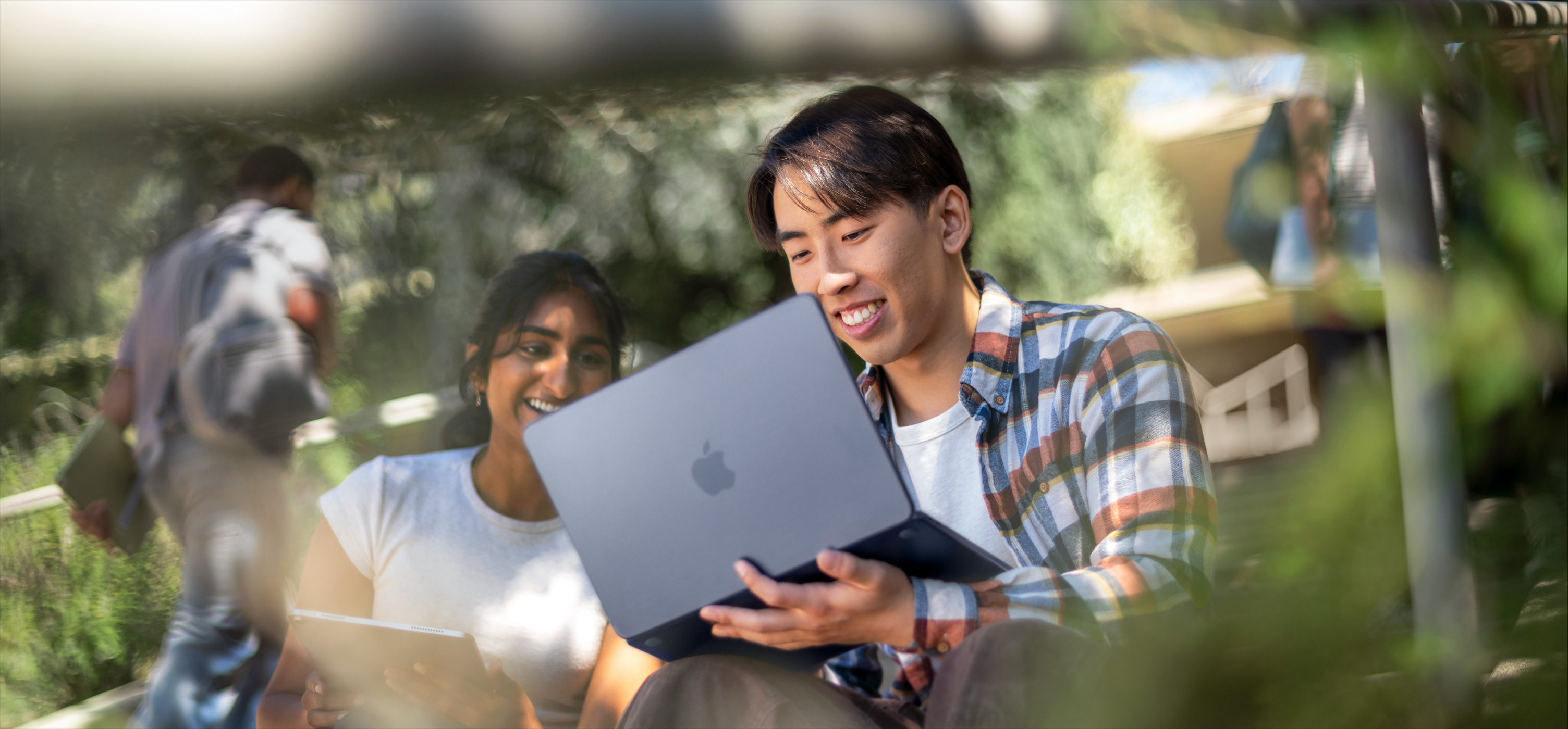 Dua siswa yang tersenyum menggunakan MacBook di luar ruangan, satu orang meletakkan MacBook itu di pangkuannya saat mereka berkolaborasi dalam sebuah tugas