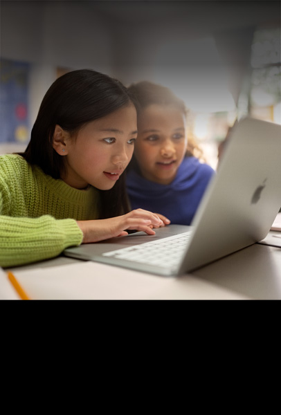 Dua siswa muda duduk di meja sambil menatap MacBook dengan penuh perhatian saat mereka berkolaborasi di kelas