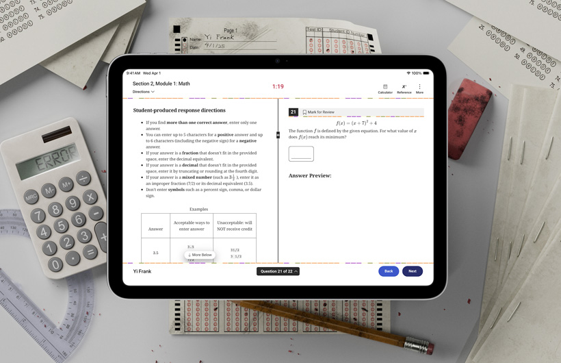An iPad sits on a desk among physical testing forms and school supplies. On screen, there are instructions for completing the math portion of a standardized test.