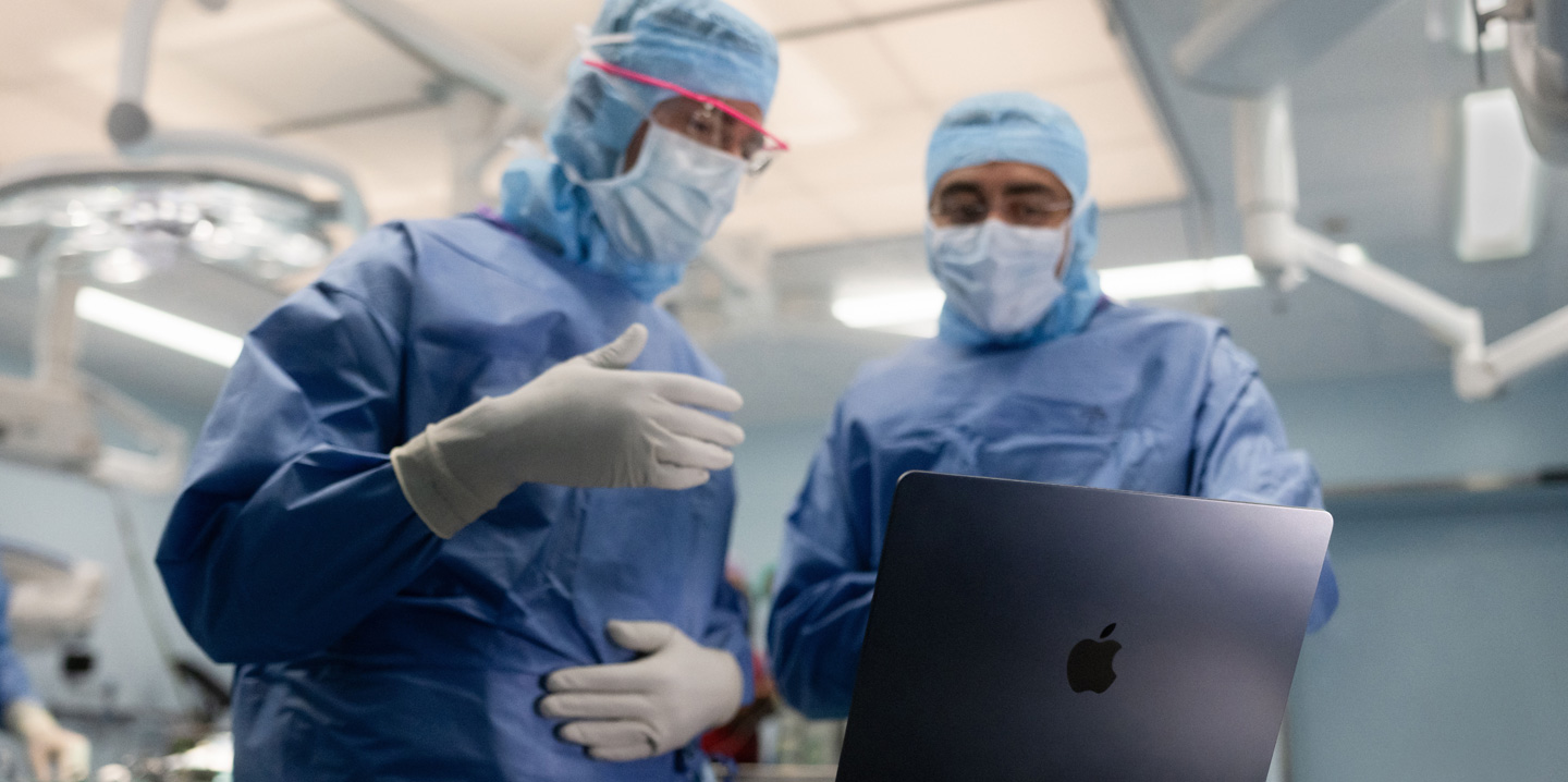 Två kirurger står i en operationssal och tittar på en MacBook. Den ena gestikulerar med handen medan den andra ser på.