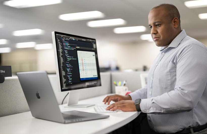 A developer stands at their desk using a Mac and MacBook with code and app information displayed on the Mac screen