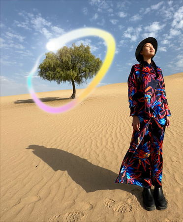 Picture of a woman in a multicoloured dress standing in a desert landscape with a lone tree in the background, circled in a glowing rainbow gradient showcasing Apple Intelligence capability to erase background objects