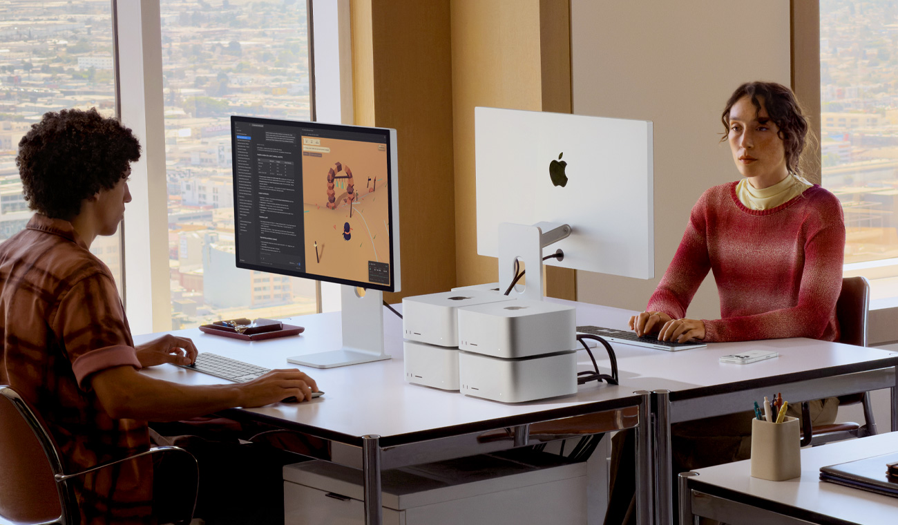 Dos personas sentadas frente a frente en sus escritorios, cuatro Mac Studio sobre un escritorio apilados de a dos, dos monitores Studio Display en los escritorios, uno muestra LM Studio y Kimi, y el otro su parte trasera