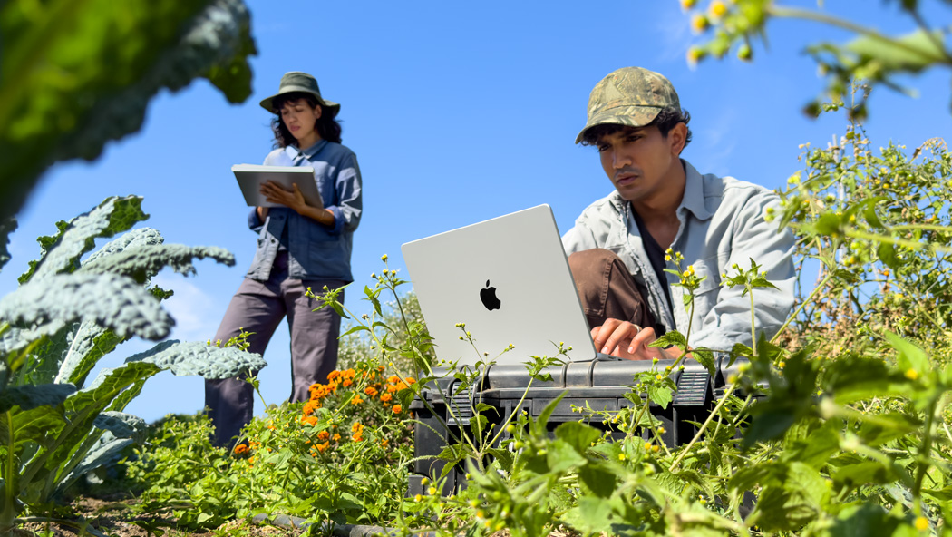 Dos personas trabajando en un jardín, una con un MacBook Pro en plata que no está enchufado y la otra con un bloc de notas