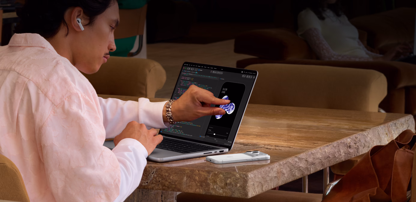 Uma pessoa sentada à mesa em um espaço público usa o gesto de pinçar para ajustar um elemento no Xcode tocando na tela de um MacBook Pro com Tela PROLED XDR prateado. Um iPhone está sobre a mesa ao lado do notebook, e a pessoa está usando os AirPods Pro