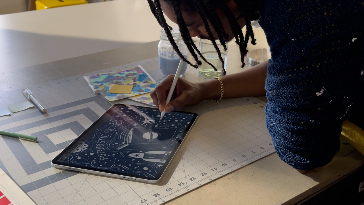 파란색 스웨터를 입고 머리를 땋은 사람이 Apple Pencil로 iPad Pro에 그림을 그리는 모습. 화면에는 행성과 우주선이 그려진 책 표지, 나비, '미래 지향적인 소설 동아리'라는 글씨가 적힌 밤하늘의 일러스트레이션이 표시되어 있습니다.