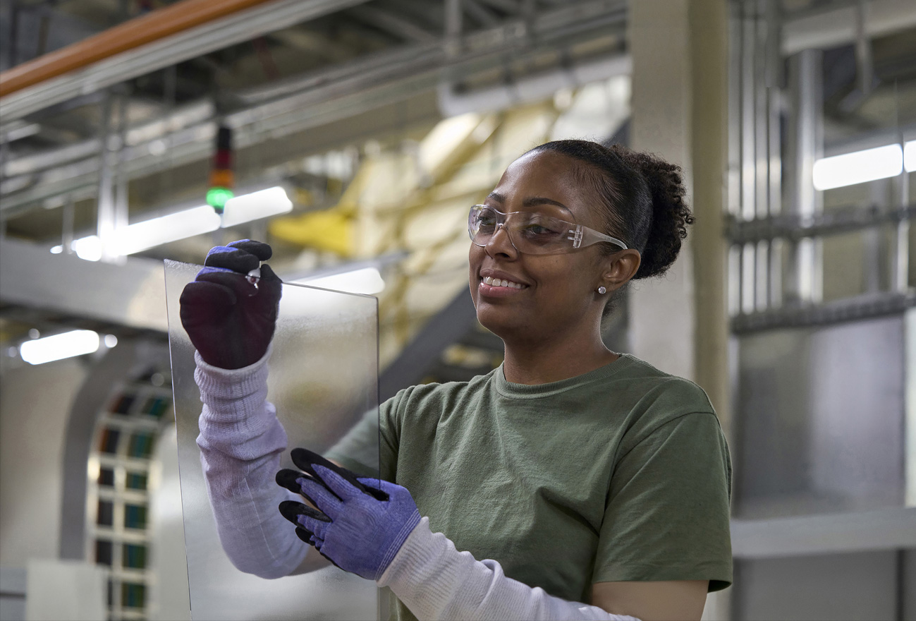 A worker wearing safety glasses inspects a glass component in an assembly facility